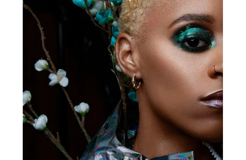 Close-up of a person's face, showing vibrant green eye makeup and glossy lips. Blonde hair and floral branches with white blossoms in the background.