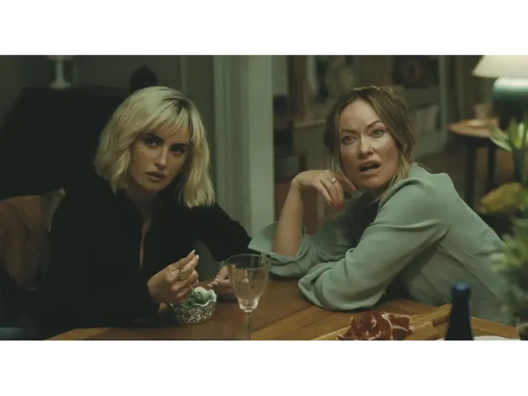 Two women sitting at a dining table, one looking surprised and the other thoughtfully eating, surrounded by food and drinks.