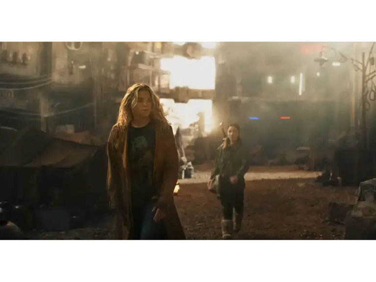 Two women walking through a desolate, urban landscape with a smoky atmosphere and makeshift tents in the background.