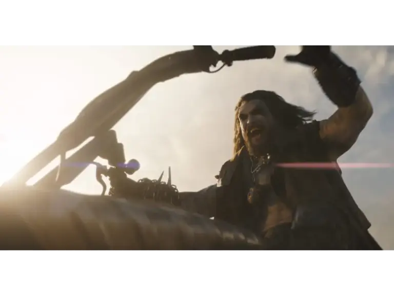 Jason Momoa as Lobo in Supergirl (2026), with long hair, is riding a motorcycle and is raising his arms enthusiastically against a bright sky.