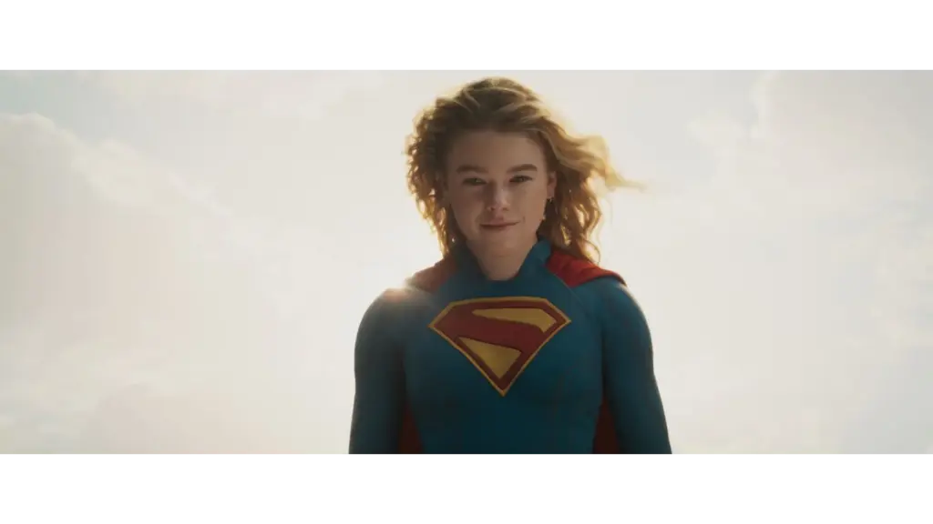 Milly Alcock in Supergirl (2026) with a blue suit and a red cape, featuring a large yellow "S" emblem on her chest, stands confidently against a bright sky.