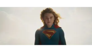 Milly Alcock in Supergirl (2026) with a blue suit and a red cape, featuring a large yellow "S" emblem on her chest, stands confidently against a bright sky.