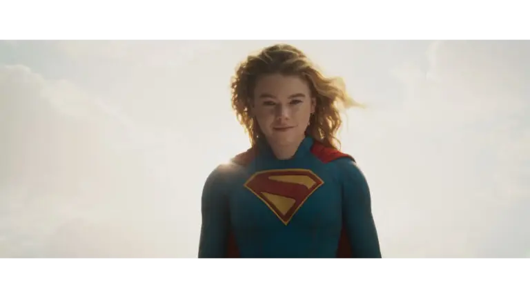 Milly Alcock in Supergirl (2026) with a blue suit and a red cape, featuring a large yellow "S" emblem on her chest, stands confidently against a bright sky.
