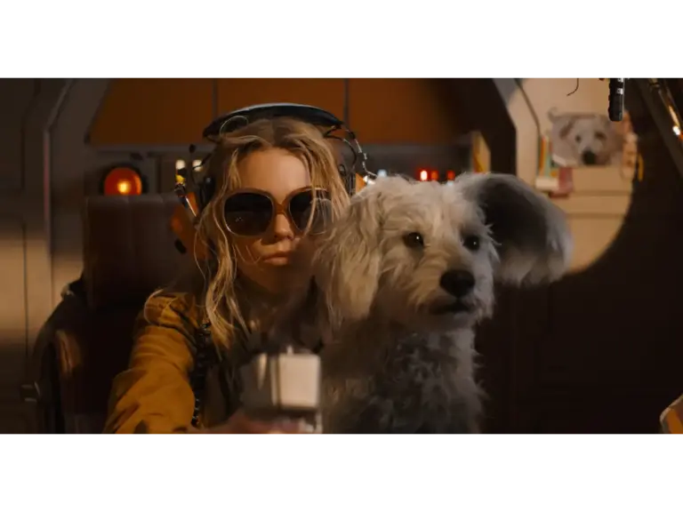 Milly Alcock in Supergirl (2026), with sunglasses and headphones, sits in a futuristic cockpit next to a fluffy dog.