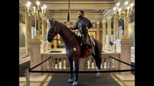 Reconstruction of a Hun warrior in full armor riding a dark horse inside a museum display, surrounded by historic decor.
