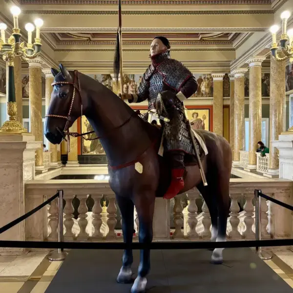 Reconstruction of a Hun warrior in full armor riding a dark horse inside a museum display, surrounded by historic decor.