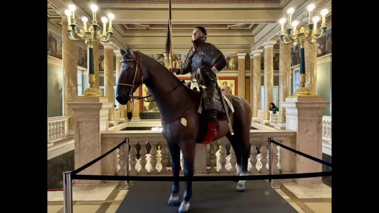 Reconstruction of a Hun warrior in full armor riding a dark horse inside a museum display, surrounded by historic decor.