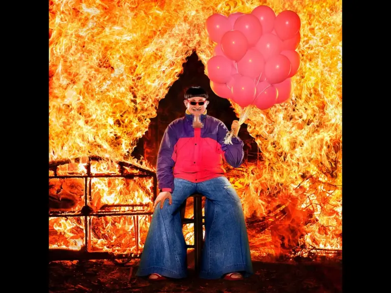 Oliver Tree sitting in front of a burning structure, holding pink balloons, wearing a purple and pink windbreaker, oversized blue JNCO Jeans, and sandals