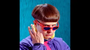 Image of Oliver Tree on a blue background, wearing orange sunglasses, a purple and yellow windbreaker, and showing the peace sign to the camera