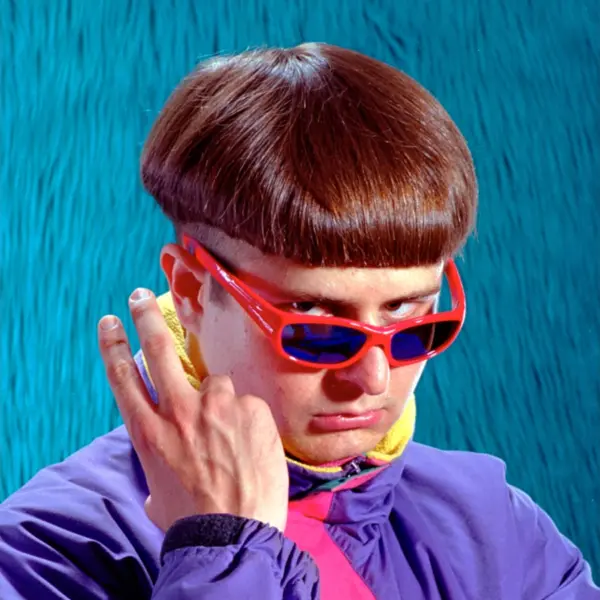 Image of Oliver Tree on a blue background, wearing orange sunglasses, a purple and yellow windbreaker, and showing the peace sign to the camera