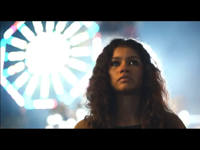 "Euphoria:" Rue Bennett thoughtfully upward in front of a bright, colorful ferris wheel at night, creating a dramatic and introspective mood.

