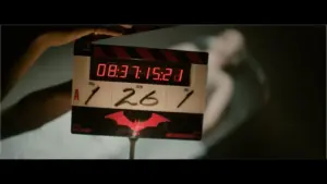 "The Batman - Part 2" - A film clapperboard with the timecode "08:37:15:23" is held in focus, featuring a red bat symbol. A blurred figure in white lies in the background.