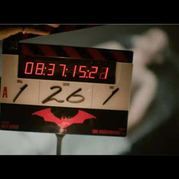 "The Batman - Part 2" - A film clapperboard with the timecode "08:37:15:23" is held in focus, featuring a red bat symbol. A blurred figure in white lies in the background.