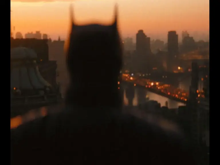 Batman over a city skyline at dusk, with warm glowing lights reflecting in a river, creating a mysterious ambiance.