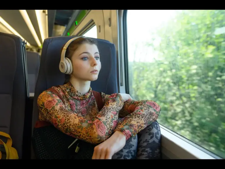 Thomasin McKenzie on a train gazes out the window, wearing headphones and a floral top, conveying a reflective and serene mood. Greenery is visible outside.