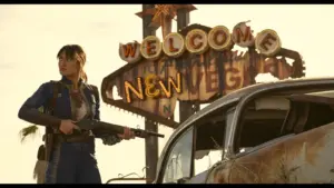 "Fallout" Lucy (Ella Purnell) stands determinedly with a shotgun in front of a worn "Welcome to New Vegas" sign. Beside her is a rusty car, evoking a post-apocalyptic scene.