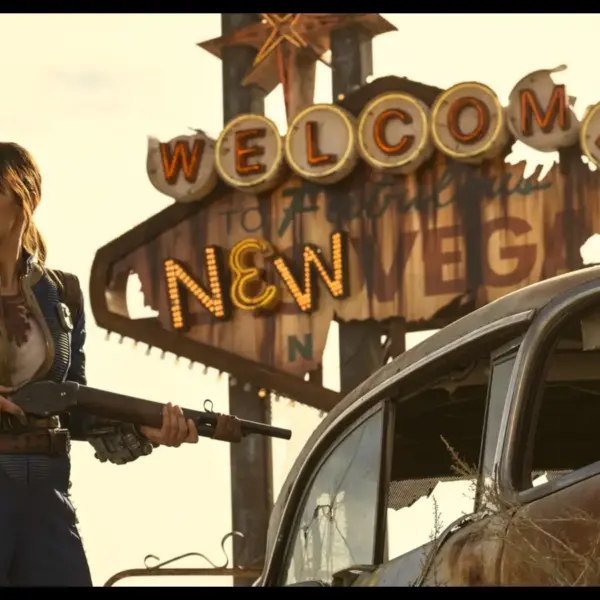 "Fallout" Lucy (Ella Purnell) stands determinedly with a shotgun in front of a worn "Welcome to New Vegas" sign. Beside her is a rusty car, evoking a post-apocalyptic scene.