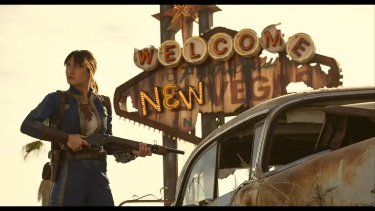 "Fallout" Lucy (Ella Purnell) stands determinedly with a shotgun in front of a worn "Welcome to New Vegas" sign. Beside her is a rusty car, evoking a post-apocalyptic scene.