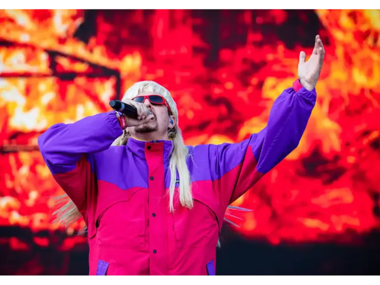 Oliver Tree, wearing a colorful jacket and sunglasses, is singing into a microphone on stage. They have long, blonde hair and are striking a dynamic pose.