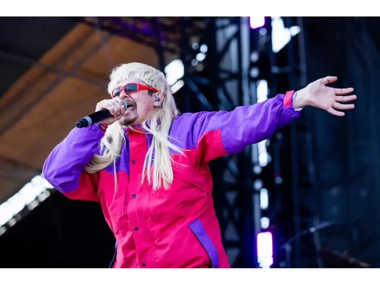 Oliver Tree is wearing a bright purple and red jacket with long blonde hair and sunglasses, singing into a microphone on stage.