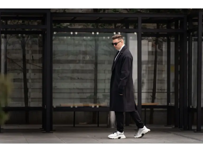 Stylish man wearing a black coat and dad sneakers stands confidently outdoors, exuding a modern fashion vibe