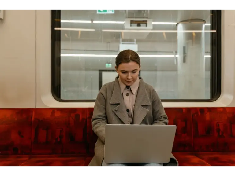 Caucasian woman using a laptop in a gray coat while sitting inside a subway train, fully immersed in her work as she embraces remote travel.