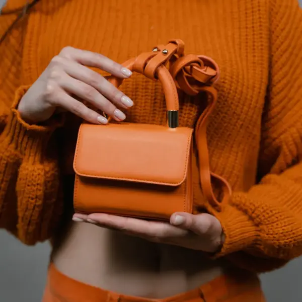 A woman holds an orange leather mini handbag against an orange sweater in a studio setting, echoing the bold charm of mini bucket bags.