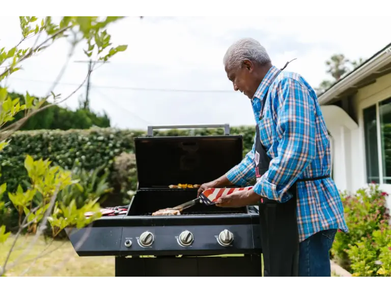 An elderly man grilling sausages on a sunny backyard afternoon, relaxing and cooking in one of his well‑used BBQ zones.