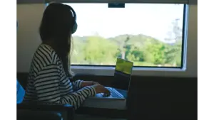 Woman using a laptop with headphones during a train journey, enjoying the scenic view while embracing the freedom of remote travel.