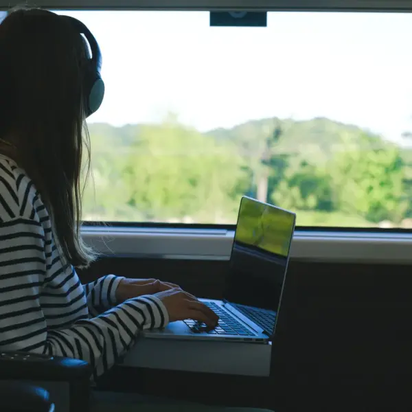 Woman using a laptop with headphones during a train journey, enjoying the scenic view while embracing the freedom of remote travel.
