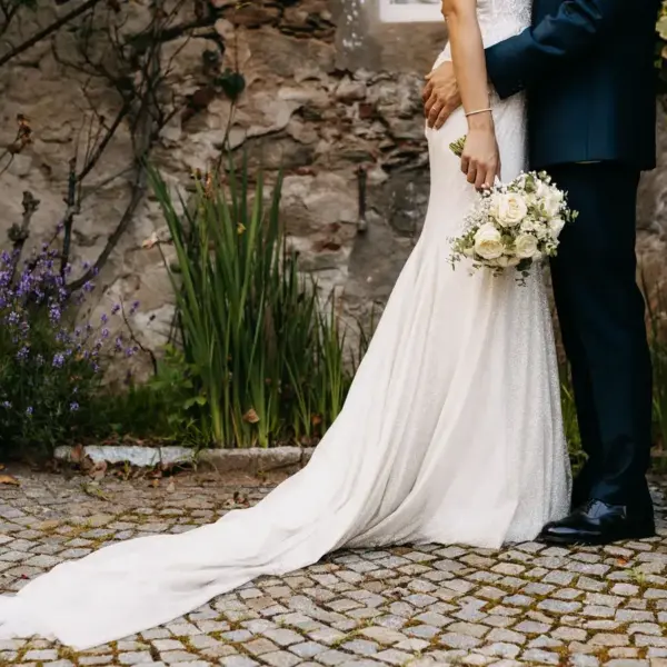 The bride in an elegant white gown holds a bouquet beside the groom in a dark suit. They stand on cobblestones with a stone wall and vibrant greenery behind. Taylor Swift and Travis Kelce's wedding rumors.