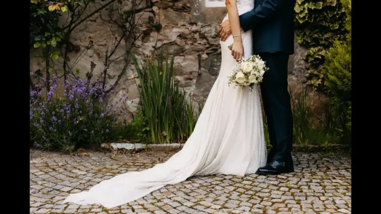 The bride in an elegant white gown holds a bouquet beside the groom in a dark suit. They stand on cobblestones with a stone wall and vibrant greenery behind. Taylor Swift and Travis Kelce's wedding rumors.
