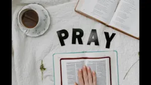 In daily devotional, someone's hand is placed on the Bible with a cup of coffee and notepad ready to study. Black letters spell out "Pray."