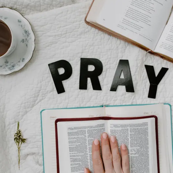 In daily devotional, someone's hand is placed on the Bible with a cup of coffee and notepad ready to study. Black letters spell out "Pray."