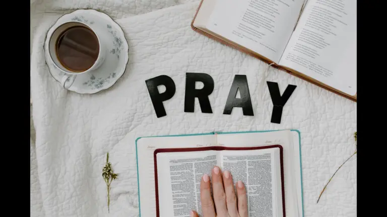 In daily devotional, someone's hand is placed on the Bible with a cup of coffee and notepad ready to study. Black letters spell out "Pray."