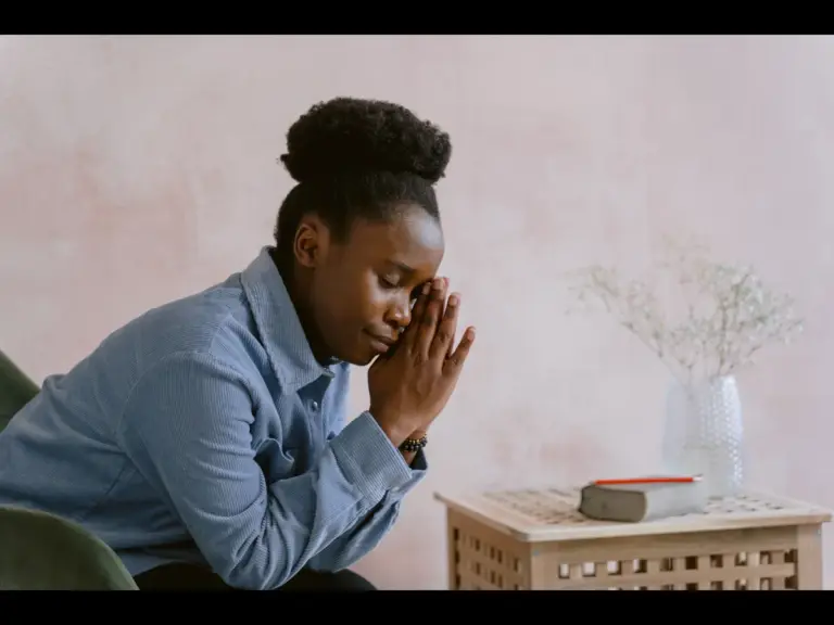In daily devotional, a woman is praying at home while sitting beside a small table.