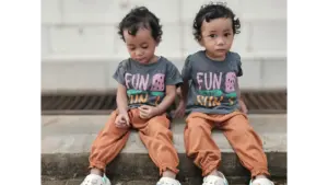 Sibling style shines through as adorable twins sit outdoors in matching outfits featuring “Fun in the Sun” T‑shirts and bright orange pants.