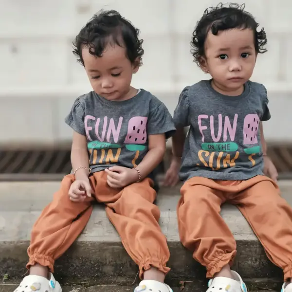 Sibling style shines through as adorable twins sit outdoors in matching outfits featuring “Fun in the Sun” T‑shirts and bright orange pants.