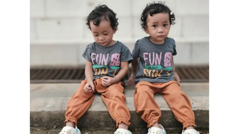 Sibling style shines through as adorable twins sit outdoors in matching outfits featuring “Fun in the Sun” T‑shirts and bright orange pants.