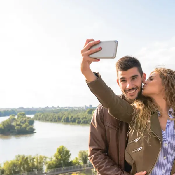 A happy couple enjoys a moment by the river, capturing a selfie to cherish their travel memories during a sweet couples travel getaway.