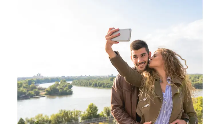 A happy couple enjoys a moment by the river, capturing a selfie to cherish their travel memories during a sweet couples travel getaway.