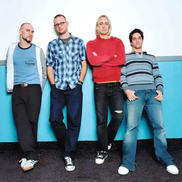 Four men, Spymob, standing side by side against a light blue wall, dressed casually in t-shirts and jeans.