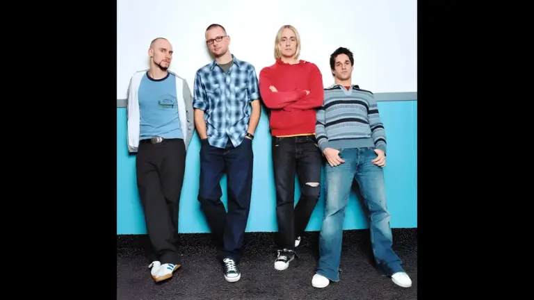 Four men, Spymob, standing side by side against a light blue wall, dressed casually in t-shirts and jeans.