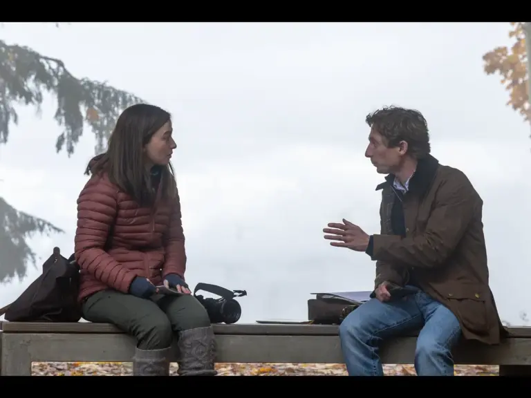 Jeremy Allen White and Mikey Madison in The Social Reckoning (2026) are sitting on a bench in a foggy outdoor setting, engaged in a conversation. The woman is wearing a red puffer jacket and the man is in a brown coat.