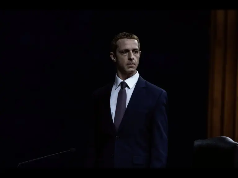 Jeremy Strong in The Social Reckoning (2026) as Mark Zuckerberg, standing in a suit, appearing before a dark backdrop, possibly in a courtroom or formal setting.