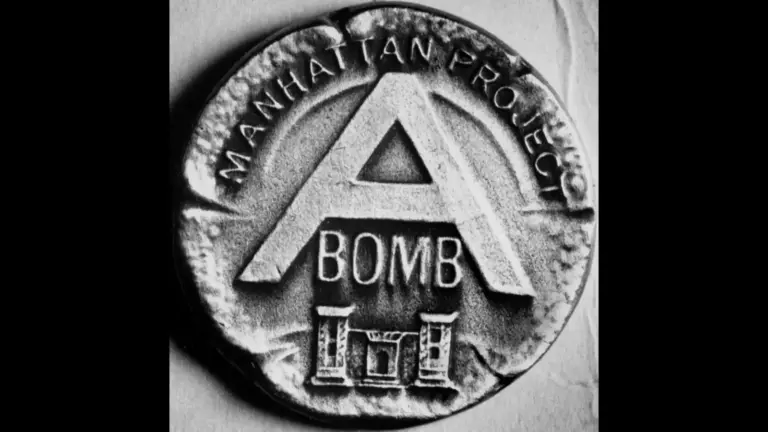 A black and white close-up photo, uploaded on July 16, showing a historical military award badge. The circular metallic pin features the letter "A" prominently in the center with the word "BOMB" underneath it. Arched along the top border of the pin are the words "MANHATTAN PROJECT."
