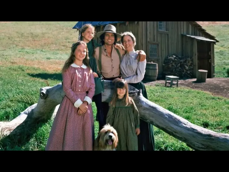 Melissa Sue Anderson, Melissa Gilbert, Michael Landon, Karen Grassle, and Sidney Greenbush in Little House on the Prairie (1974), standing together outdoors near a log with a house and grassy hill in the background. A dog is seated in front of them.