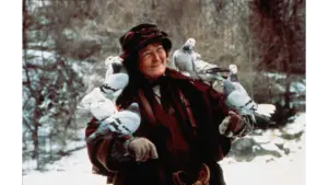 Brenda Fricker in Home Alone 2: Lost in New York (1992) wearing a dark coat and hat, smiling, with several pigeons perched on her arms and shoulders in a snowy outdoor setting.