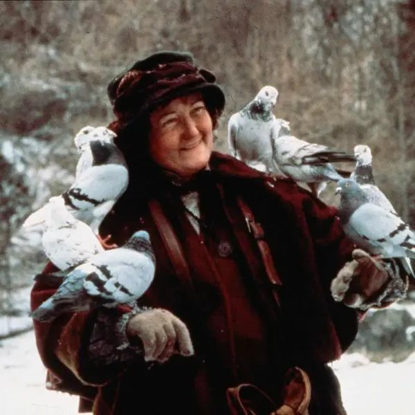 Brenda Fricker in Home Alone 2: Lost in New York (1992) wearing a dark coat and hat, smiling, with several pigeons perched on her arms and shoulders in a snowy outdoor setting.