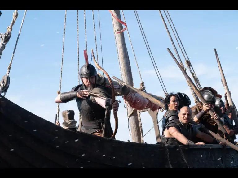 A promotional still from the 2026 film "The Odyssey" featuring Matt Damon, Will Yun Lee, and Jimmy Gonzales aboard a wooden vessel under a bright, clear blue sky. On the left, Matt Damon stands near the bow, leaning forward with his arms braced against the wooden structure. He wears a dark, metallic helmet with a prominent red crest and dark leather armor. Further back on the right, several other crew members, including Will Yun Lee and Jimmy Gonzales, look out across the water. The ship's complex network of thick ropes, wooden rigging, and a large mast frames the background.
